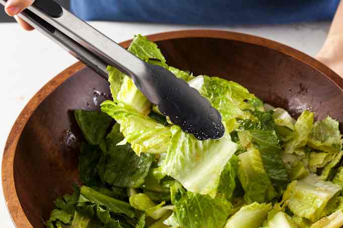 Pair of tongs being used to toss pieces of lettuce in a wooden salad bowl.